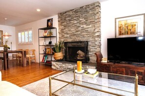 A living room with a stone fireplace, a flat-screen TV, a glass coffee table, and a bookshelf.