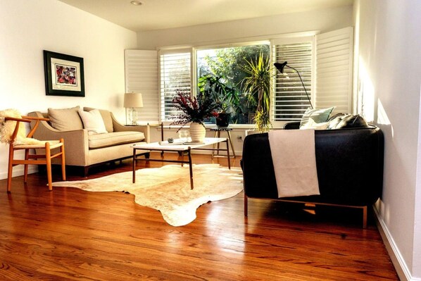 A living room with a sofa, a chair, a coffee table, and a rug. There are windows with blinds, a lamp, and a plant.