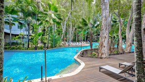 Une piscine entourée de palmiers et de chaises longues, avec une terrasse en bois et un panneau indiquant qu’il est interdit de se baigner.