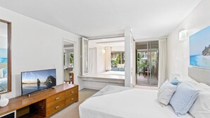 Een hotelkamer met een bed, een televisie op een houten dressoir, een balkon met uitzicht op de oceaan en een schilderij van een strand.