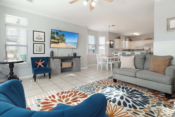 A spacious living room with a large sofa, a blue armchair, and a colorful rug. There is a dining area with a table and chairs, and a kitchen with white cabinets and appliances.