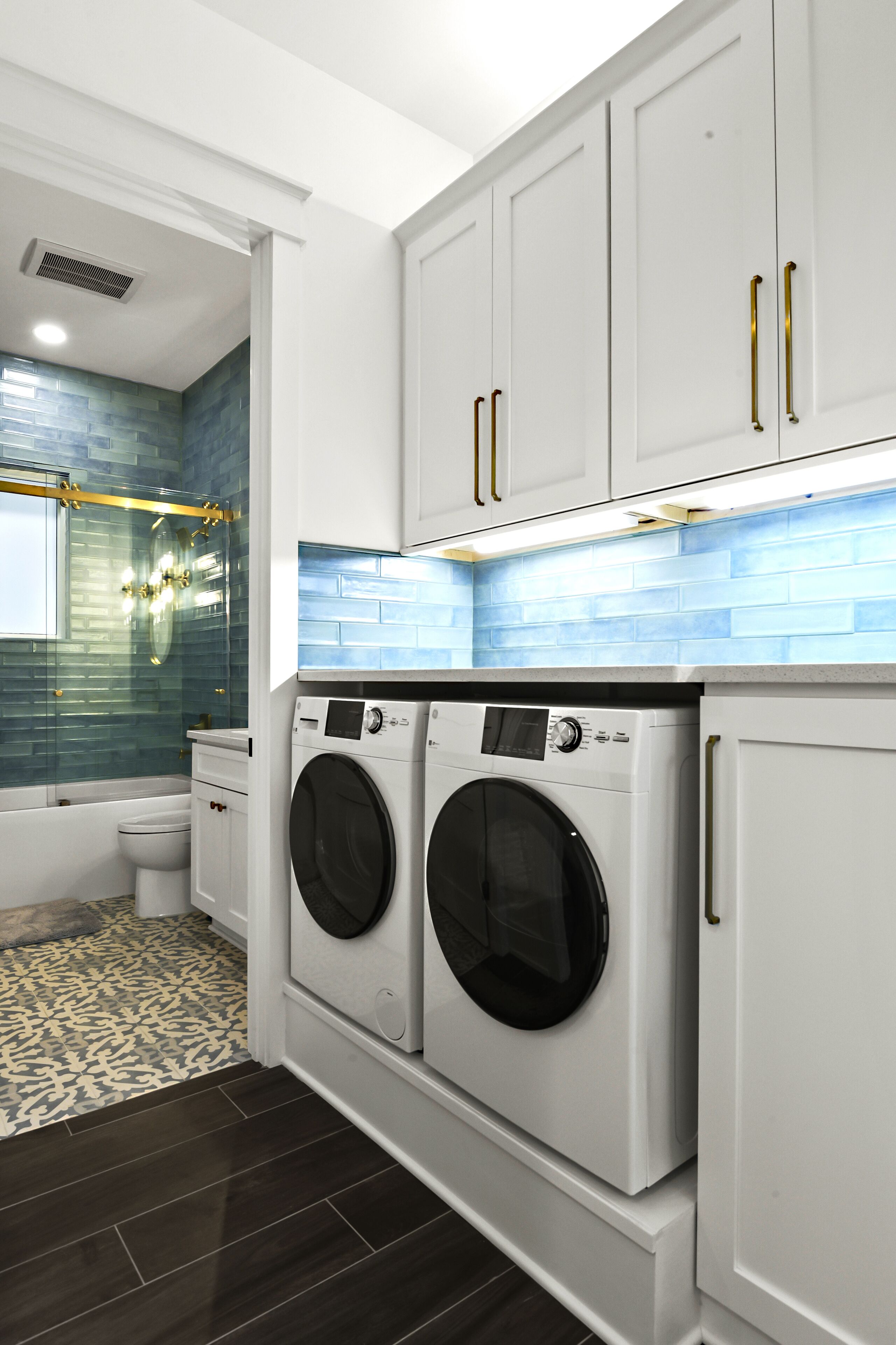 A modern laundry room with two front-loading washing machines, a white toilet, and a blue-tiled shower area.