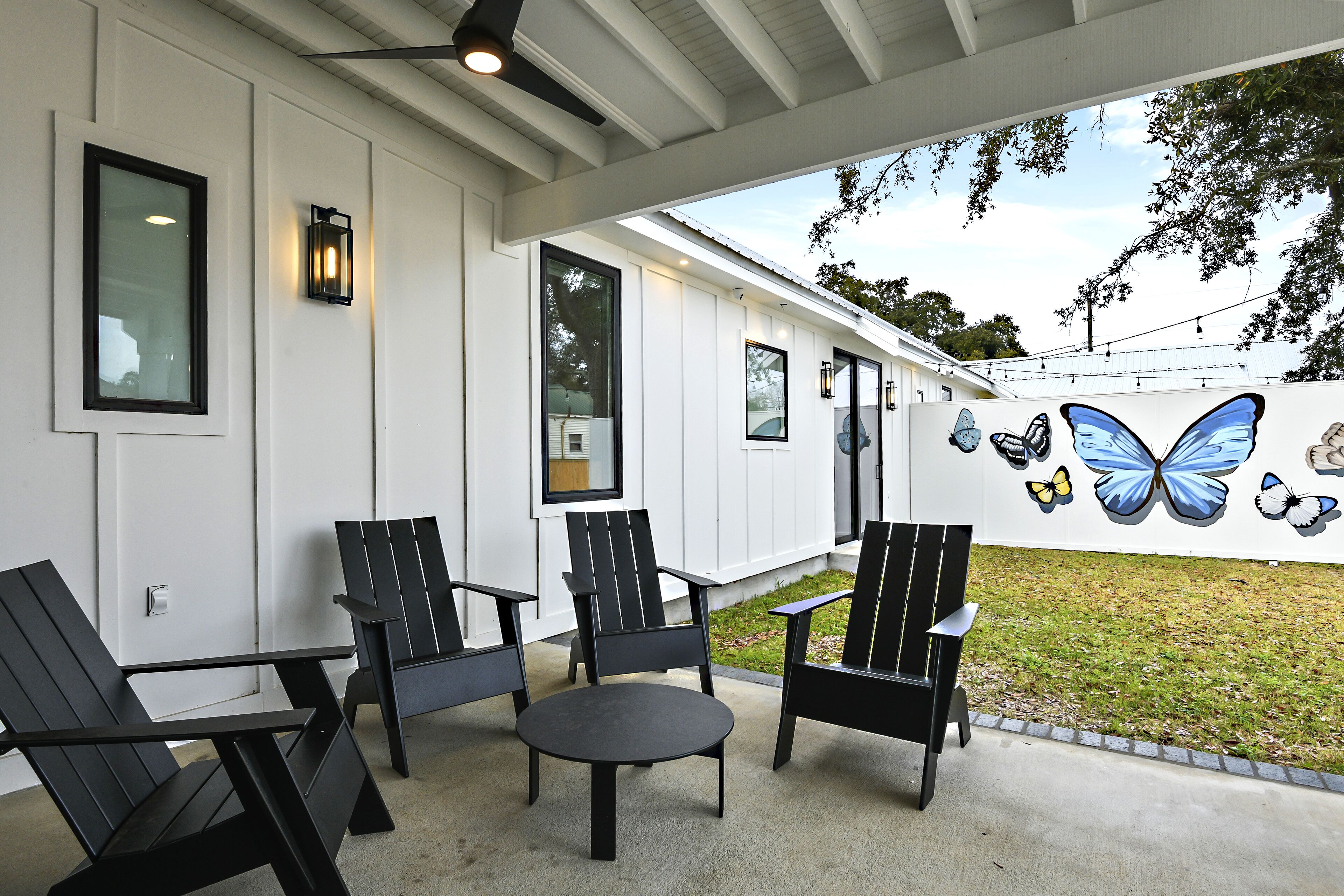 Une terrasse couverte avec quatre chaises Adirondack noires, une petite table ronde et une fresque murale représentant des papillons.