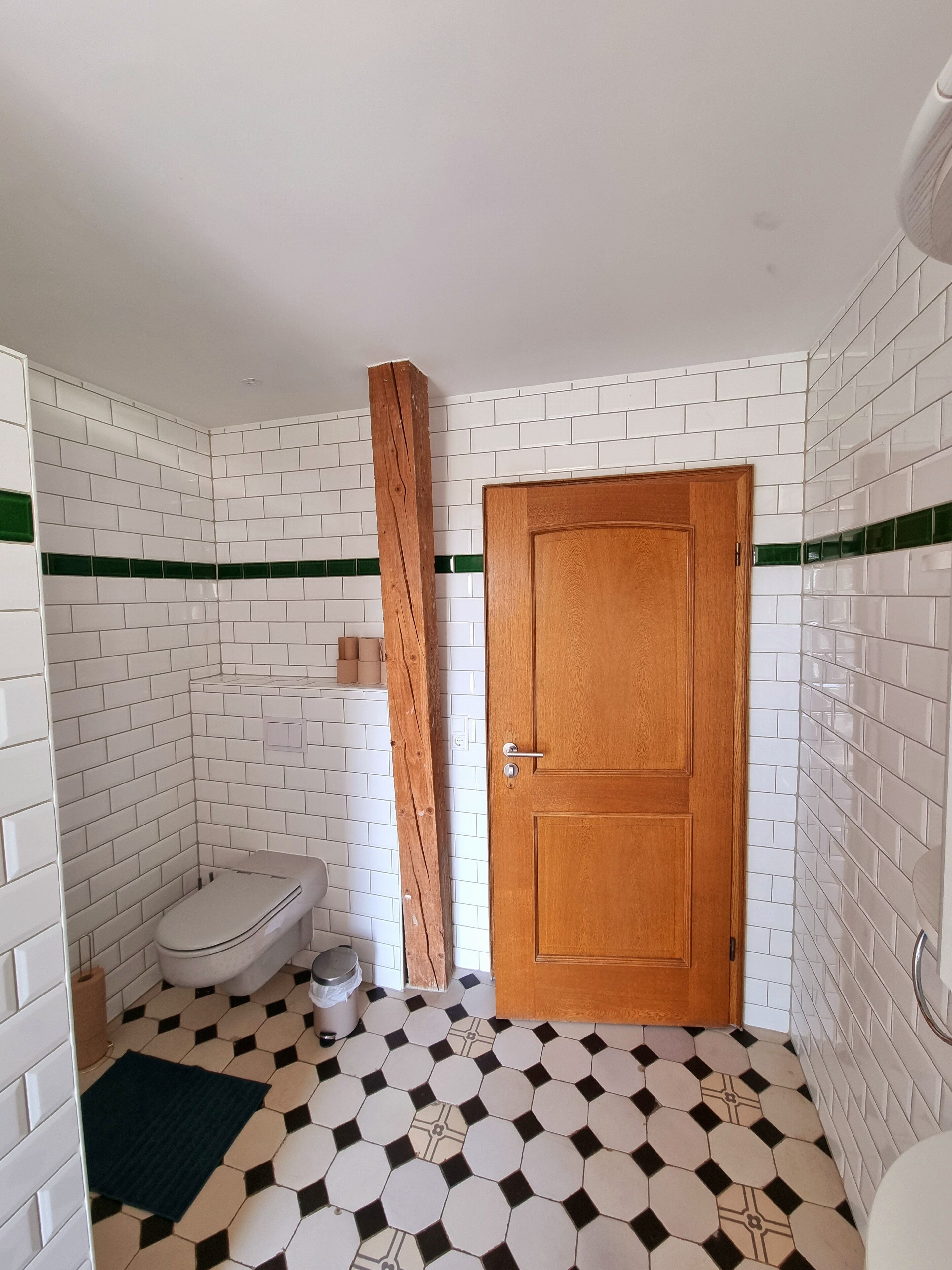 Un baño con baldosas hexagonales blanco y negro, una puerta de madera, un inodoro y una franja de baldosas a rayas verdes.