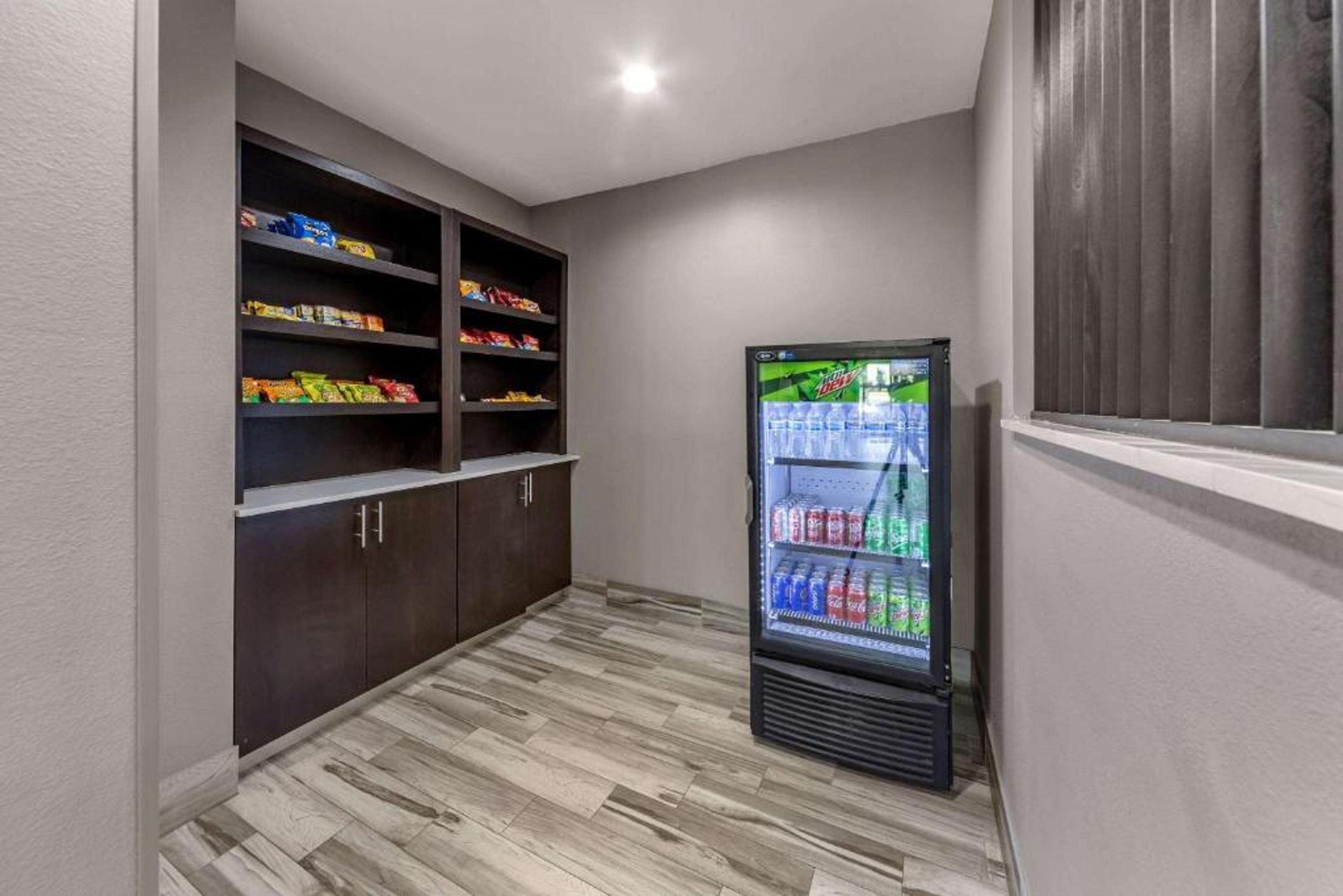 A narrow hallway with a refrigerator stocked with beverages, shelves with snacks, and a window with blinds.