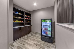 Un pasillo estrecho con un refrigerador lleno de bebidas, estantes con aperitivos y una ventana con persianas.