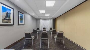 A conference room with rows of chairs, a white curtain, framed pictures on the wall, and a carpeted floor.