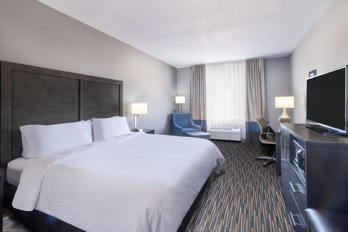 Quarto de hotel com uma cama grande, duas mesas de cabeceira, uma escrivaninha, uma cadeira, uma televisão e uma janela com cortinas.