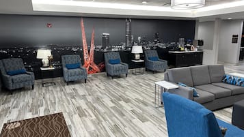 A modern hotel lobby with a cityscape view, grey patterned chairs, a grey sofa, and a bar area.