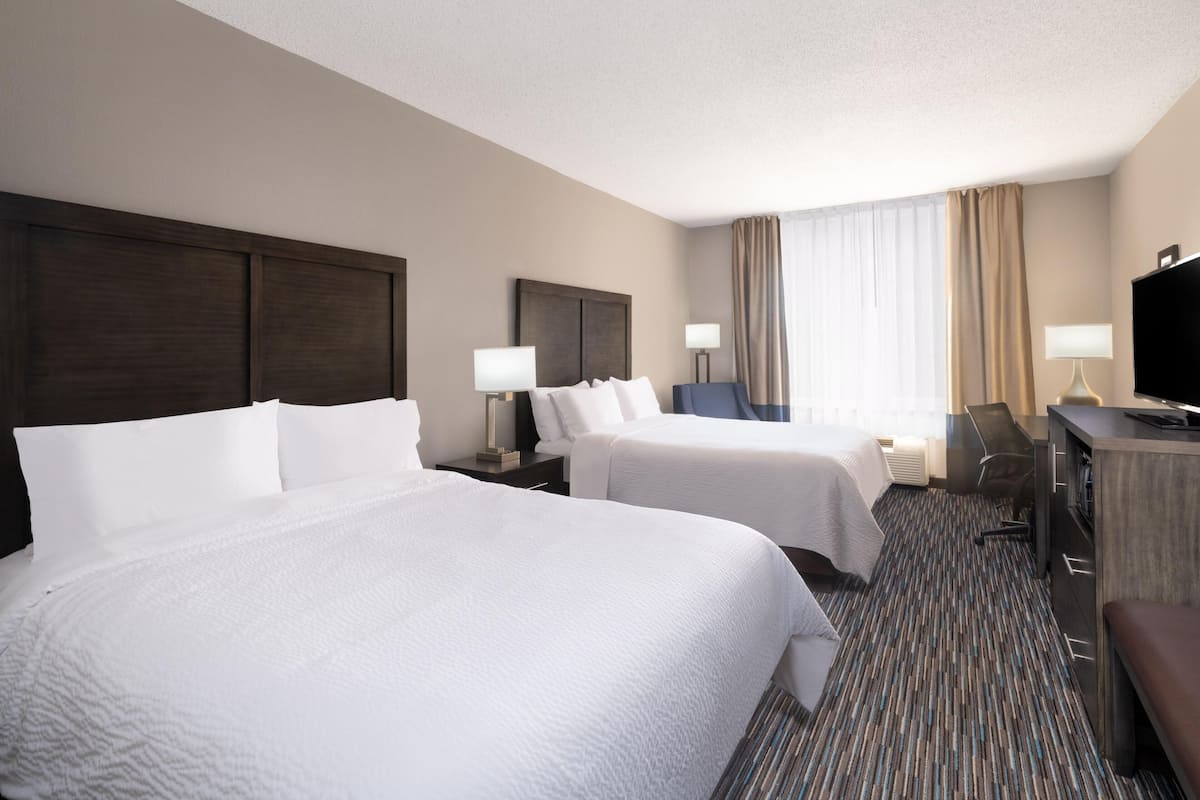 Quarto de hotel com duas camas, uma escrivaninha, uma cadeira, televisão e janela com cortinas.