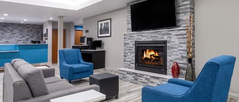 A modern living room with a fireplace, blue and gray sofas, a flat-screen TV, and a kitchen area in the background.