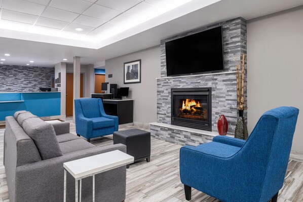 A modern living room with a fireplace, blue and gray sofas, a flat-screen TV, and a kitchen area in the background.