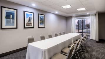 A conference room with a long table, chairs, framed pictures on the wall, and a large window with curtains.
