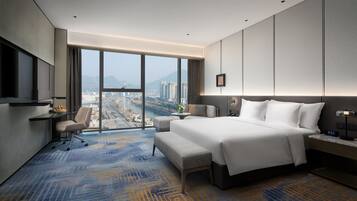 A modern hotel room with a large bed, a desk, a chair, and a view of a cityscape through large windows.