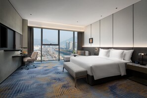 A modern hotel room with a large bed, a desk, a chair, and a view of a cityscape through large windows. - Crowne Plaza Shangluo (Shanglou)