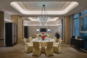 Un comedor moderno con una mesa redonda central dispuesta para seis personas, rodeada de sillas color beige. Un gran candelabro cuelga del techo, y un televisor de pantalla plana está montado en la pared.