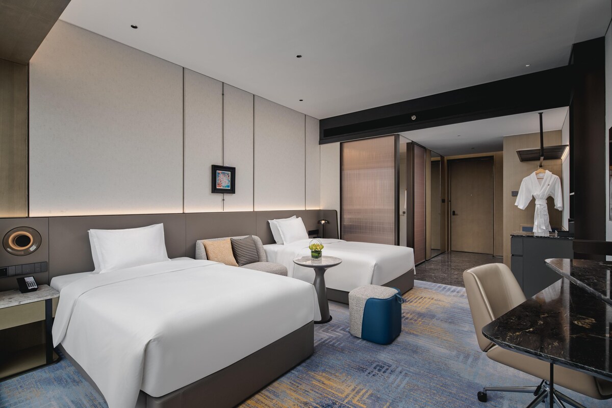 A modern hotel room with two beds, a small round table, a desk with a phone, and a seating area with a chair.