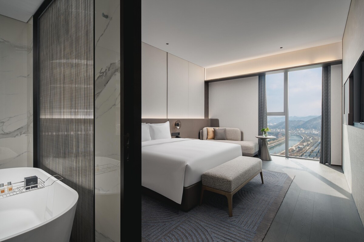 Una habitación de hotel moderna con una cama grande, una zona de estar, vistas a un paisaje urbano y un baño con una bañera espaciosa.