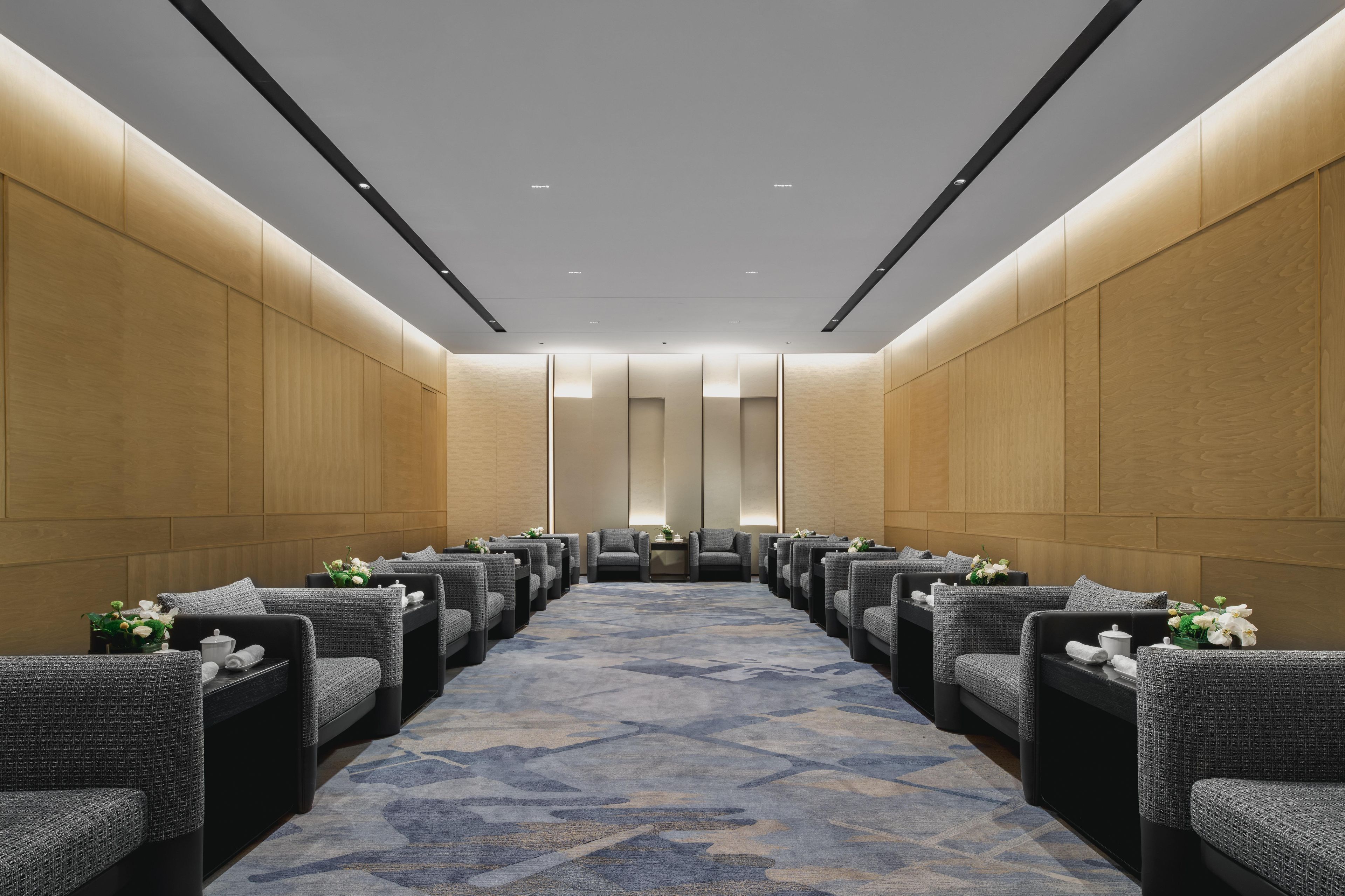 A hotel room with a long corridor, grey upholstered chairs, a patterned carpet, and wooden panel walls.