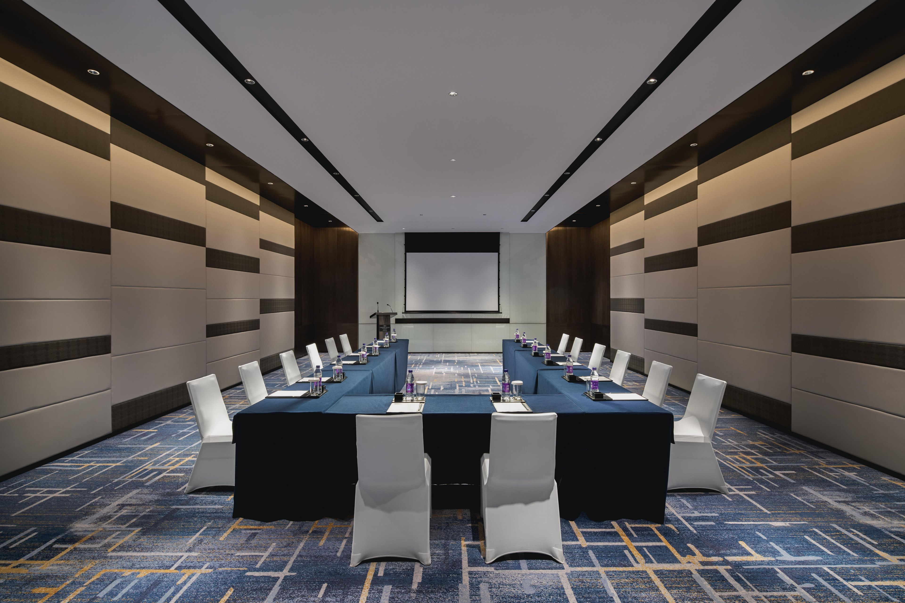 A conference room with a long table, white chairs, and a projector screen.