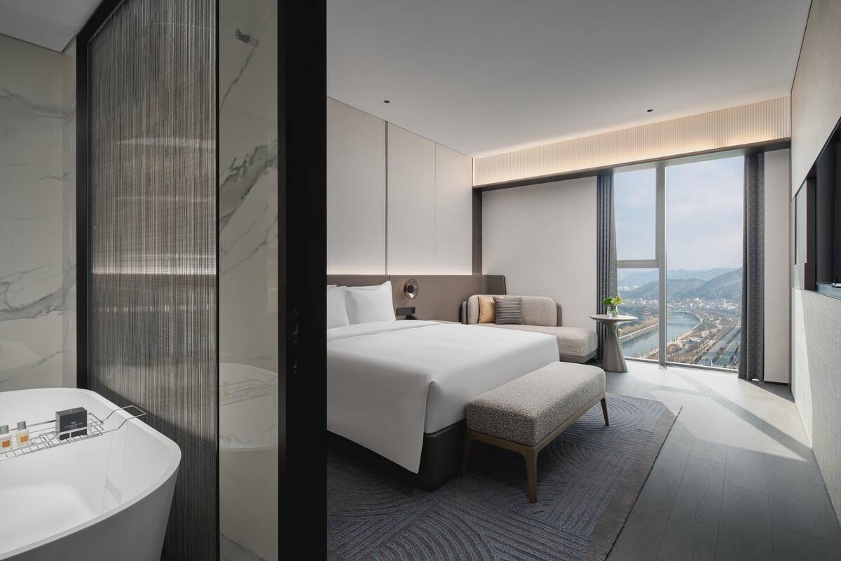 A modern hotel room with a large bed, a sitting area, a view of a cityscape, and a bathroom with a large bathtub.