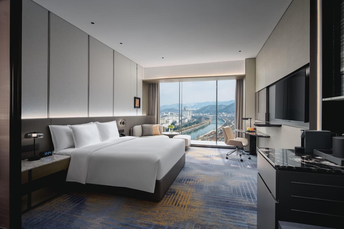 A modern hotel room with a large bed, a desk, a TV, and a view of a city and mountains.