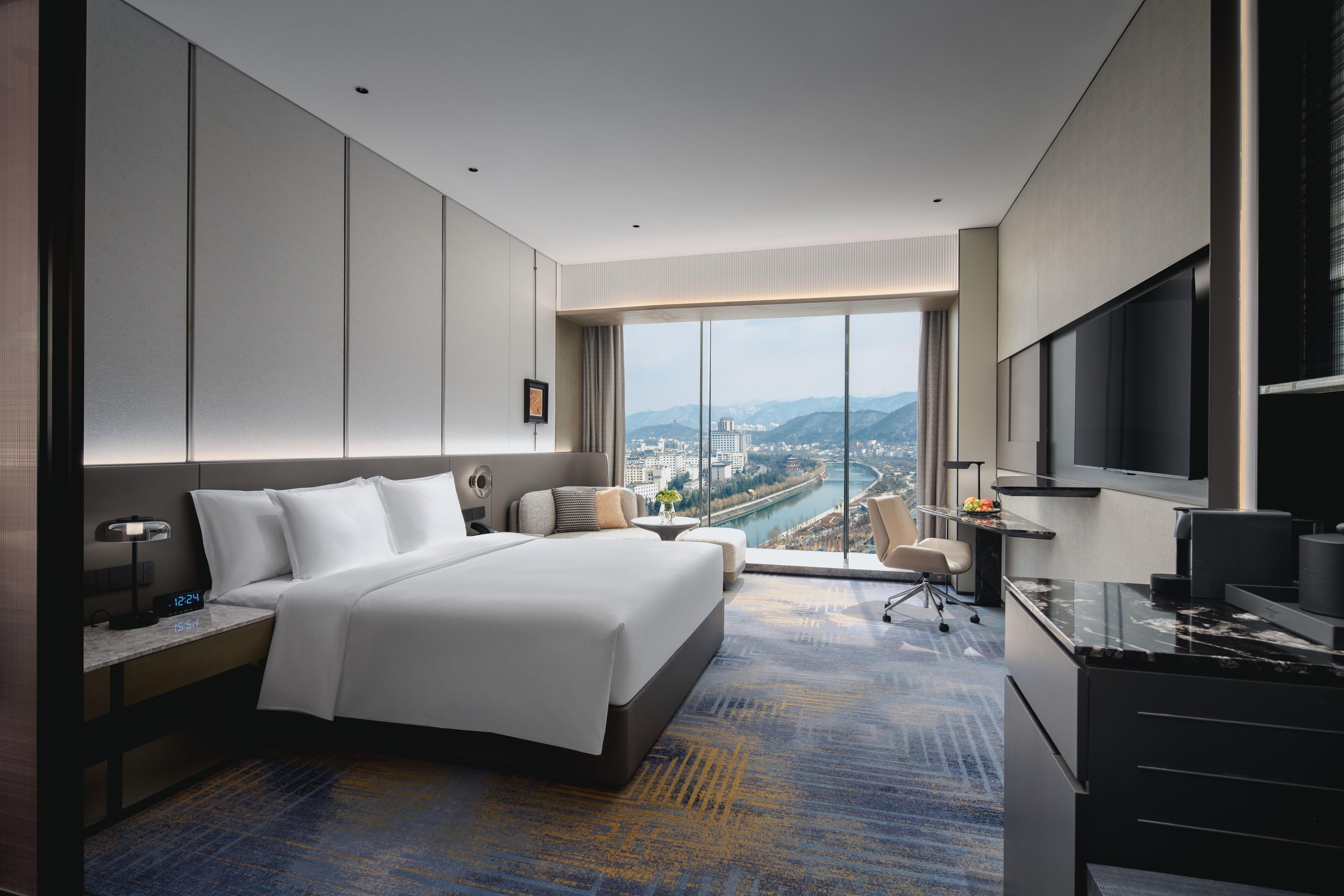 Una habitación de hotel moderna con una cama grande, un escritorio, un televisor y vistas a una ciudad y montañas.