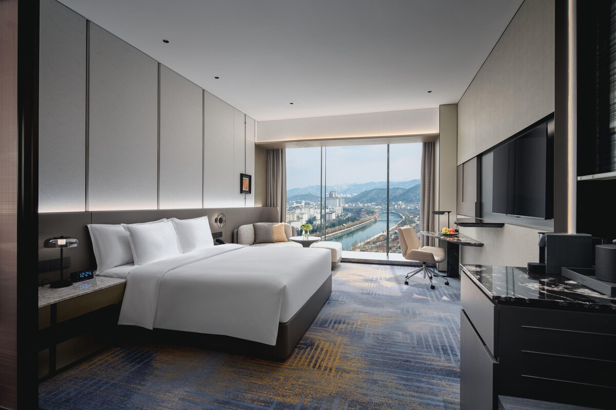 Una habitación de hotel moderna con una cama grande, un escritorio, un televisor y vistas a una ciudad y montañas.