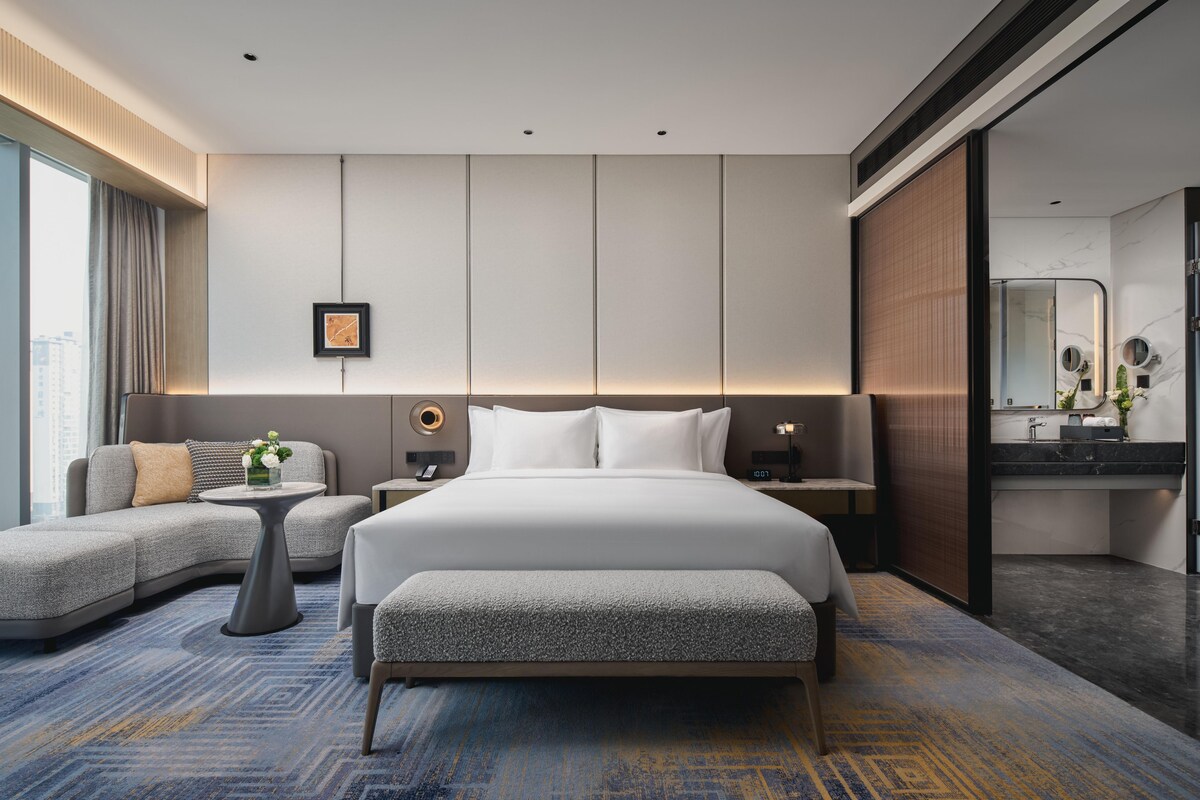 Una habitación de hotel moderna con una cama grande, un sofá, una mesita y un baño con espejo.