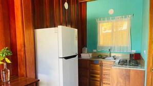 A compact kitchen with a white refrigerator, wooden cabinets, a gas stove, and a window with sheer curtains. - Charming lodge in beautiful Playa Samara with WiFi (Playa Samara Guanacaste Province)