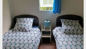 A room with two beds, each with patterned bedding, a small table with a roll of toilet paper, and a window with a blue curtain. - Mobilhome Ty Fregamano, Terrain Privé, vue mer (Locquirec)