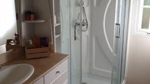 A bathroom with a glass-enclosed shower, a white sink, and a mirror above it. There are shelves with potted plants and toiletries. - Mobilhome Ty Fregamano, Terrain Privé, vue mer (Locquirec)