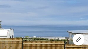 A view of a coastal area with a wooden fence, a white trailer, and a building with a metal roof. - Mobilhome Ty Fregamano, Terrain Privé, vue mer (Locquirec)