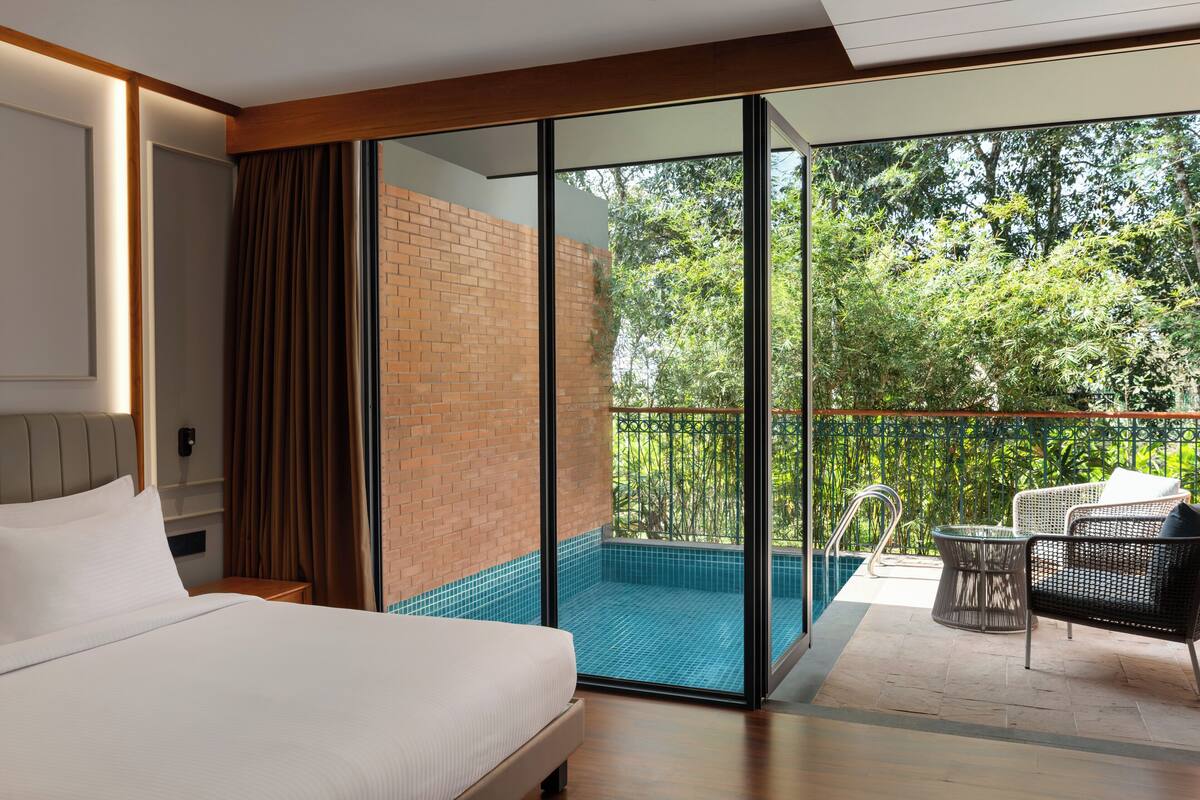A hotel room with a bed, a glass door leading to a balcony with a pool and outdoor furniture.