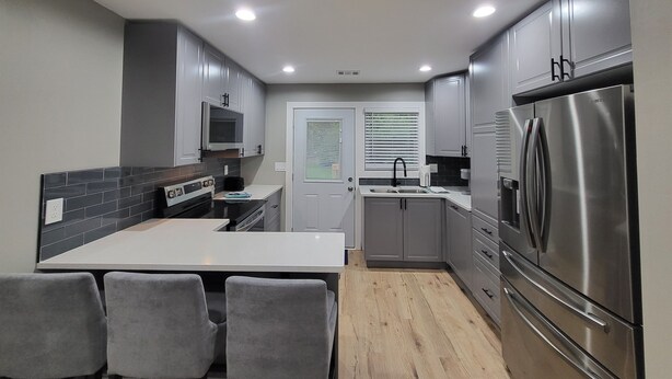A modern kitchen with grey cabinets, a stainless steel refrigerator, a built-in oven, and a dining area with grey chairs.