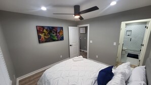 A bedroom with a bed, a ceiling fan, a colorful abstract painting, and a bathroom visible through an open door.
