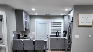 A modern kitchen with grey cabinets, a dining area with two chairs, a ceiling fan, recessed lighting, and a coffee table with a remote control.