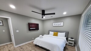 Um quarto com cama, ventilador de teto, televisão fixada na parede e janela com persianas.