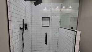 Um chuveiro com paredes de vidro, revestido com azulejos hexagonais brancos e cinzas, um porta-toalhas preto e um pequeno espelho quadrado na parede.