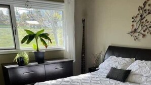 A bedroom with a large bed, a nightstand, a dresser, a window with a view of greenery, and a wall-mounted plant. - Charming 2 bedroom home.🏔 (Cranbrook)