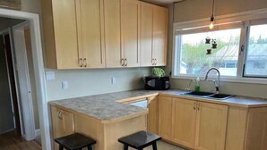 A kitchen with wooden cabinets, a granite countertop, two stools, a sink, and a window with natural light. - Charming 2 bedroom home.🏔 (Cranbrook)