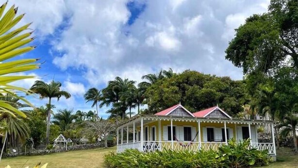 A two-story house with a red roof, surrounded by lush greenery and palm trees.