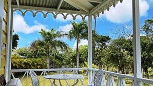 A covered outdoor seating area with a table and two chairs, surrounded by a white railing and overlooking a lush garden with palm trees.