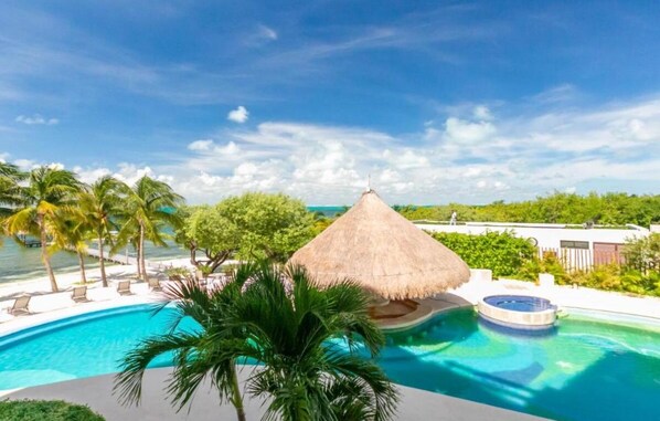A pool area with a thatched-roof structure, palm trees, and a clear view of the ocean.