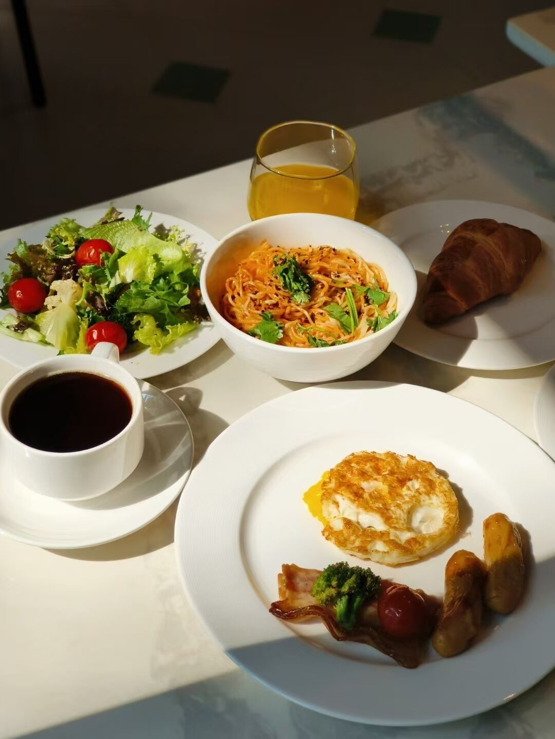 A table with a meal consisting of a salad, noodles, a croissant, an omelette, and a cup of coffee.