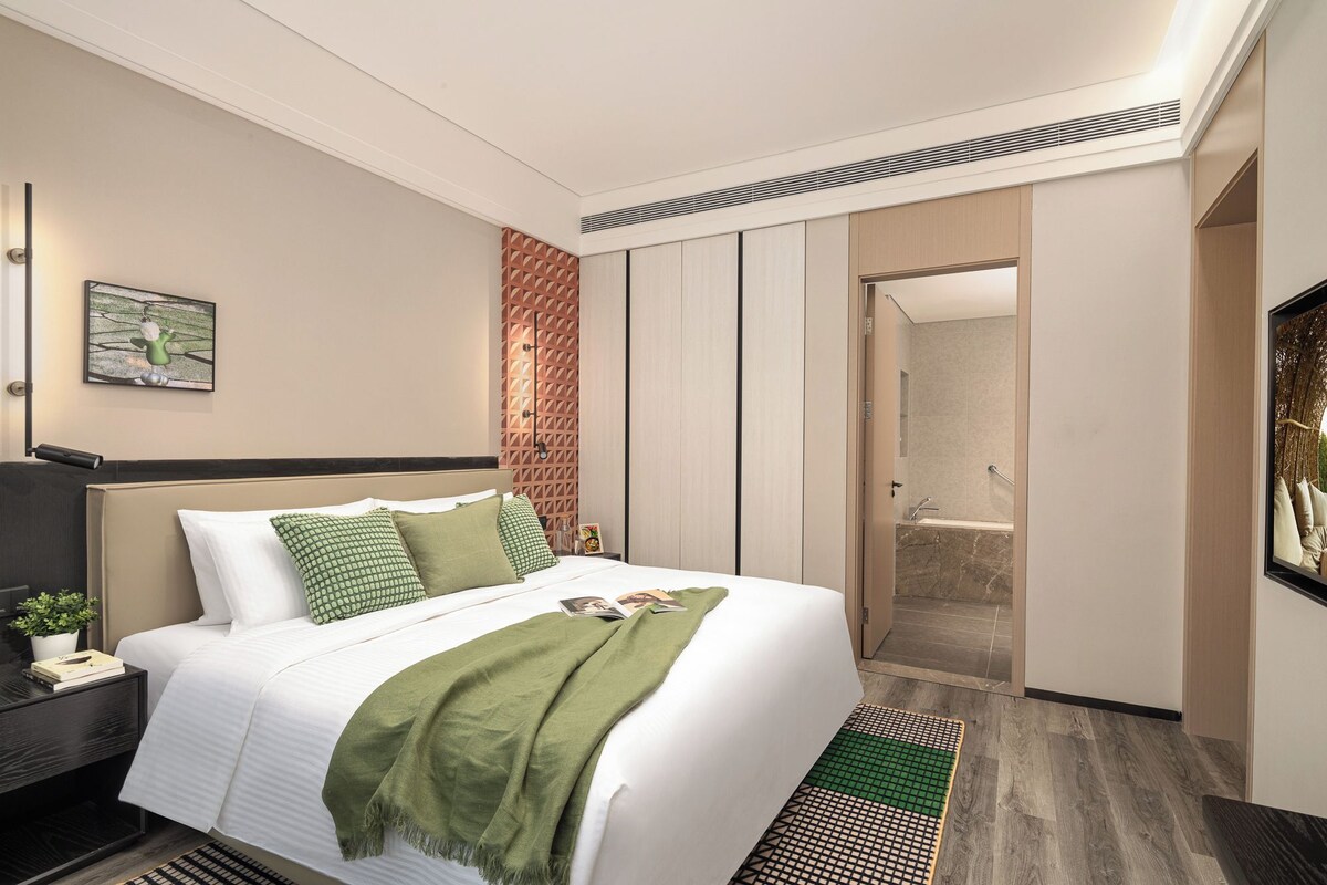 Une chambre d’hôtel moderne avec un grand lit, une table de chevet avec une lampe, un plaid vert et une vue sur la salle de bain.