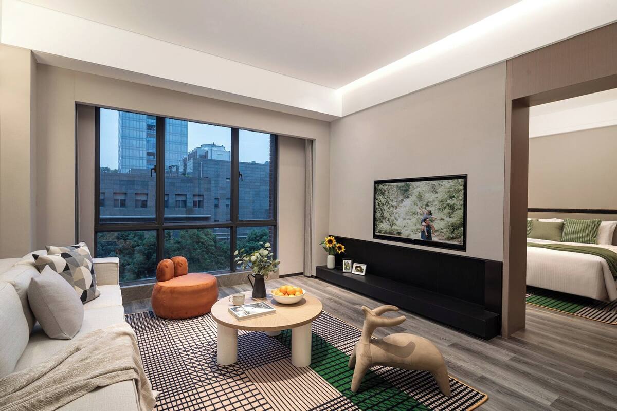 Een moderne hotelkamer met een groot raam, een bank, een bijzettafeltje met een boek en bloemen, een televisie en een bed met een groen-witte dekbedovertrek.