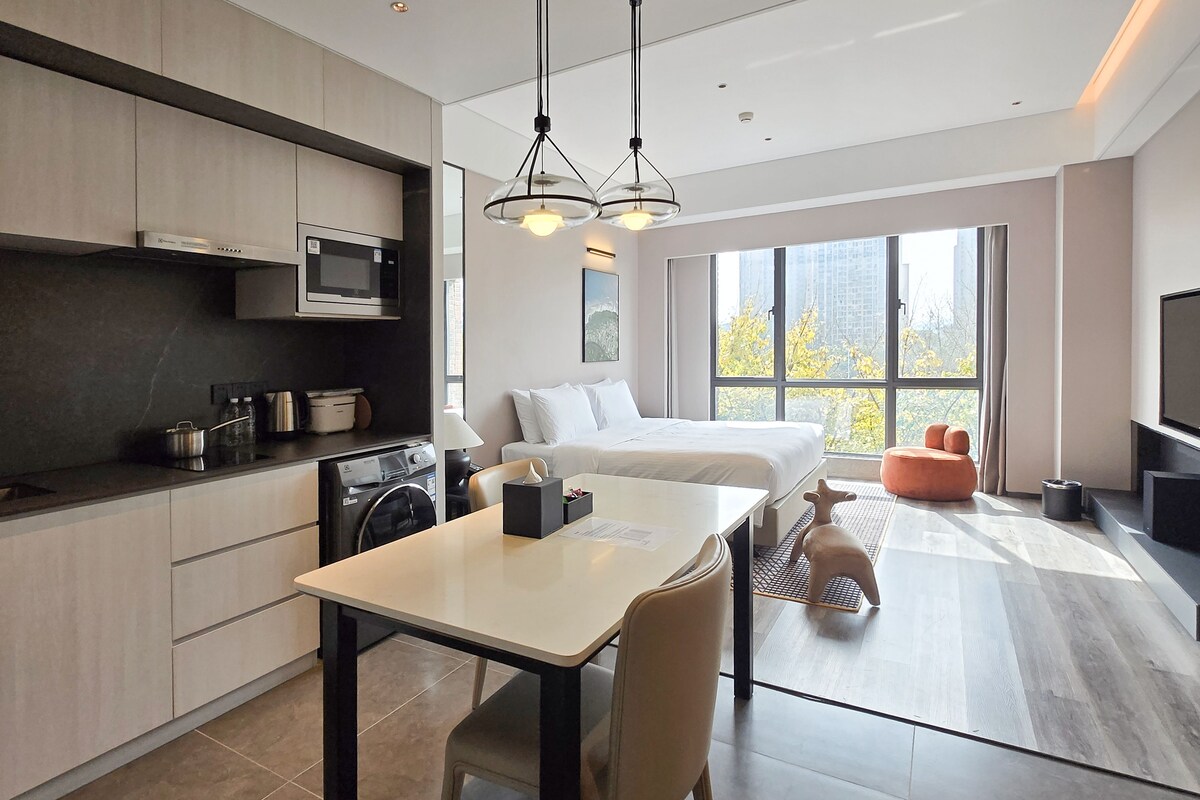 Une chambre d’hôtel moderne dotée d’une cuisine, d’un coin repas, d’un lit et d’une grande fenêtre donnant sur la ville.