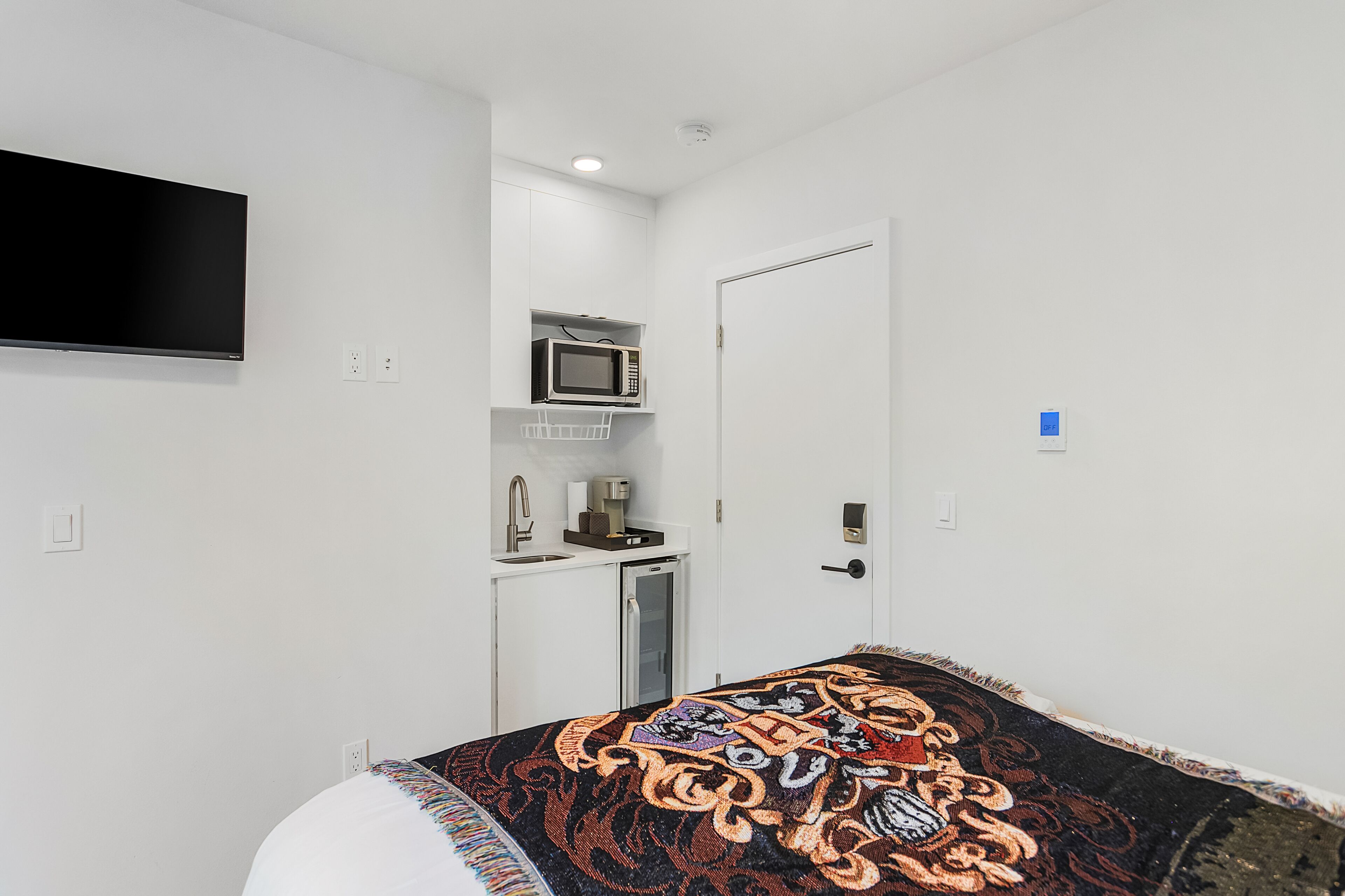 Une chambre d’hôtel équipée d’un lit, d’un téléviseur à écran plat fixé au mur, d’un four à micro-ondes et d’un petit coin cuisine.