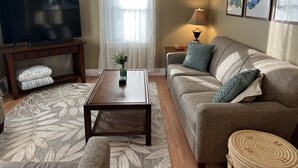 A living room with a television on a wooden stand, a sofa with patterned cushions, a coffee table with a vase, and a lamp on a side table. - Off season weekly and monthly rates! 700 weekly, 2400 monthly (Muskegon)